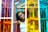A woman stands with her arms outstretched inside a structure made of multicolored glass panels creating a vibrant and dynamic environment. Her expression is serene and slightly pensive as sunlight illuminates her face and the translucent colors cast intricate patterns of light and shadow.