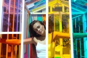 A woman stands with her arms outstretched inside a structure made of multicolored glass panels creating a vibrant and dynamic environment. Her expression is serene and slightly pensive as sunlight illuminates her face and the translucent colors cast intricate patterns of light and shadow.