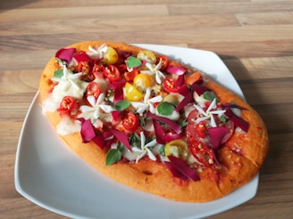 A colorful flatbread served on a white plate is topped with fresh ingredients. There are cherry tomatoes, both red and yellow, slices of cheese, and green leaves. Red and pink flower petals are also scattered across the surface, adding a decorative and unusual touch.