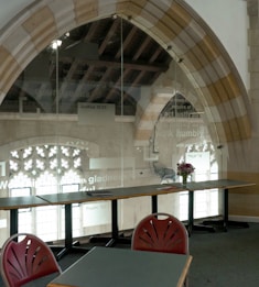 A room with arched windows and glass panels displaying biblical verses. Tables and red chairs are arranged in a row, and a flower vase sits on one table. The architecture features a blend of stone and glass, with ambient light from outside illuminating the space.