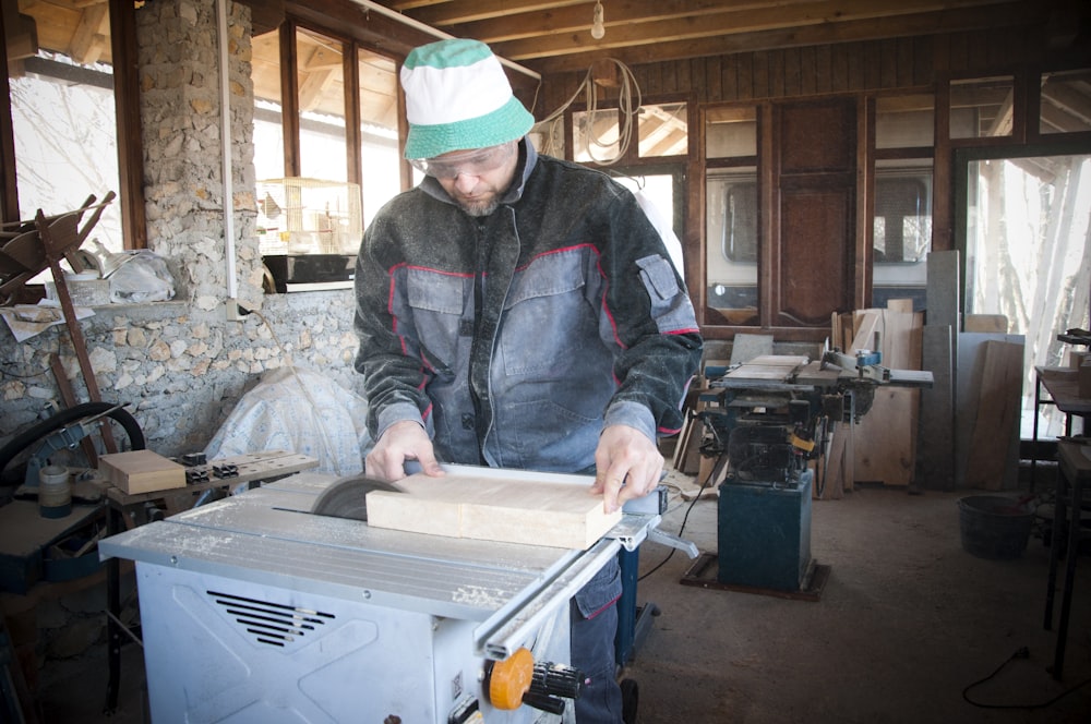 Persona che indossa giacca nera e grigia che taglia una tavola di legno sulla sega da tavolo