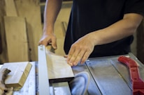Hands guiding a wooden plank through a table saw in a workshop setting, surrounded by various woodworking tools.
