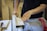 Hands guiding a wooden plank through a table saw in a workshop setting, surrounded by various woodworking tools.