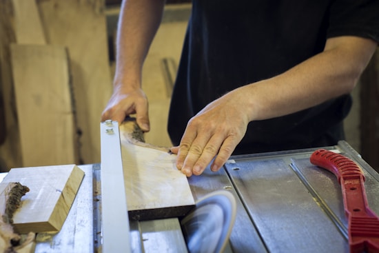Hands guiding a wooden plank through a table saw in a workshop setting, surrounded by various woodworking tools.