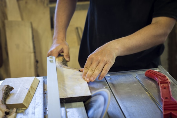 Hands guiding a wooden plank through a table saw in a workshop setting, surrounded by various woodworking tools.