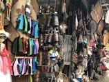 A lively and colorful market stall filled with various handcrafted items and decorations. Crocheted garments in vibrant hues of blue, pink, green, and red hang on display. Copper pots, hanging bells, and ropes are neatly arranged, creating an atmosphere rich with texture and cultural significance. A doll adorned in a pink dress stands prominently on the left side. Numerous other traditional crafts and trinkets fill the space, creating a sense of abundance and eclecticism.