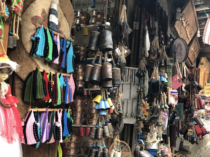 A lively and colorful market stall filled with various handcrafted items and decorations. Crocheted garments in vibrant hues of blue, pink, green, and red hang on display. Copper pots, hanging bells, and ropes are neatly arranged, creating an atmosphere rich with texture and cultural significance. A doll adorned in a pink dress stands prominently on the left side. Numerous other traditional crafts and trinkets fill the space, creating a sense of abundance and eclecticism.