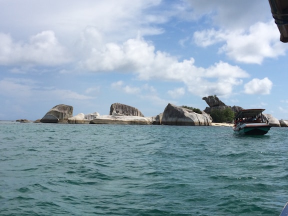 Rock formations rise from the turquoise sea under a partly cloudy sky. A boat with an awning carries several people near the rocky shore, suggesting a tour or excursion.