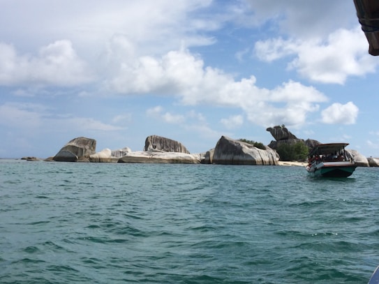 Rock formations rise from the turquoise sea under a partly cloudy sky. A boat with an awning carries several people near the rocky shore, suggesting a tour or excursion.