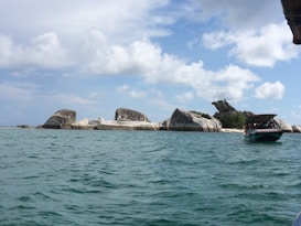 Rock formations rise from the turquoise sea under a partly cloudy sky. A boat with an awning carries several people near the rocky shore, suggesting a tour or excursion.
