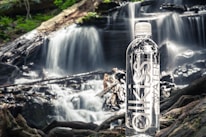 A bottle of water stands prominently on a piece of wood in front of a cascading waterfall. The surroundings are lush with greenery, and the water flows over rocks and fallen branches, creating a serene natural scene.
