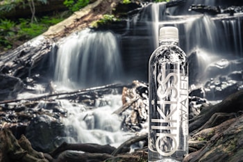 A bottle of water stands prominently on a piece of wood in front of a cascading waterfall. The surroundings are lush with greenery, and the water flows over rocks and fallen branches, creating a serene natural scene.