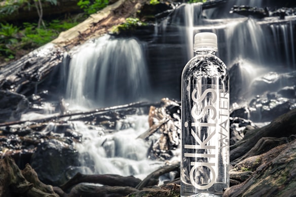 A bottle of water stands prominently on a piece of wood in front of a cascading waterfall. The surroundings are lush with greenery, and the water flows over rocks and fallen branches, creating a serene natural scene.