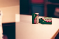 Two boxes of Fujicolor C200 film are positioned on a white surface. One box is upright, and the other lies flat. The background is blurry, displaying shades that hint at a room interior.