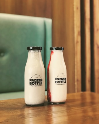 Two glass bottles of milkshakes are placed on a wooden table, with a logo that reads 'Frozen Bottle' on the front. One bottle is plain white, while the other has a red swirled pattern. The background includes a tufted, soft teal seating area and wooden wall panels.