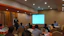 A workshop session is taking place in a conference room. Several people are seated around tables with water bottles in front of them, while a speaker stands near a whiteboard and speaks to the audience. A large screen displays a presentation or document, and another person sits near the screen. The room is well-lit and the atmosphere appears professional.