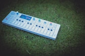 A synthesizer with multiple buttons and knobs is placed on green grass. The control panel includes a small display screen and a series of keys similar to a piano. The lively colors on the device contrast against the natural backdrop.