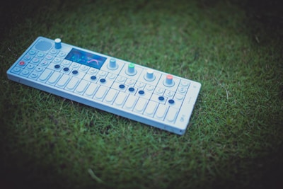A synthesizer with multiple buttons and knobs is placed on green grass. The control panel includes a small display screen and a series of keys similar to a piano. The lively colors on the device contrast against the natural backdrop.