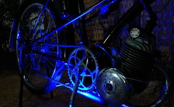 A close-up of a bicycle's mechanical components, featuring a chain, gear, and a metallic engine part illuminated with blue lighting. The background consists of a woven texture, possibly a fence.