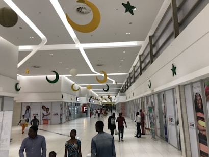 A spacious shopping mall corridor featuring a high ceiling with modern lighting. Decorative elements such as crescent moons and stars hang from the ceiling, adding a festive atmosphere. Several people are walking and interacting, some carrying shopping bags.