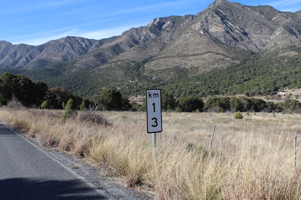 A scenic landscape with a road on the left leading into the distance. A kilometer marker sign indicating 13 kilometers is visible in the foreground, surrounded by dry grasses. The background features a range of tall, rugged mountains covered in patches of green vegetation against a clear blue sky.