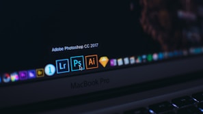 A close-up of a MacBook Pro laptop screen displaying several application icons on the dock. The highlighted application is Adobe Photoshop CC 2017, accompanied by other software icons like Lightroom and Illustrator. The cursor is pointing at the Photoshop icon.
