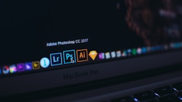 A close-up of a MacBook Pro laptop screen displaying several application icons on the dock. The highlighted application is Adobe Photoshop CC 2017, accompanied by other software icons like Lightroom and Illustrator. The cursor is pointing at the Photoshop icon.