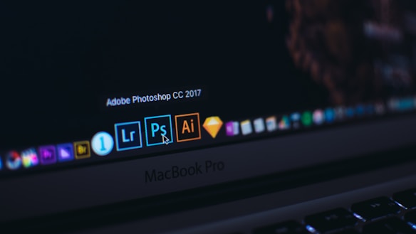 A close-up of a MacBook Pro laptop screen displaying several application icons on the dock. The highlighted application is Adobe Photoshop CC 2017, accompanied by other software icons like Lightroom and Illustrator. The cursor is pointing at the Photoshop icon.
