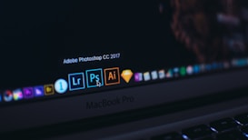 A close-up of a MacBook Pro laptop screen displaying several application icons on the dock. The highlighted application is Adobe Photoshop CC 2017, accompanied by other software icons like Lightroom and Illustrator. The cursor is pointing at the Photoshop icon.