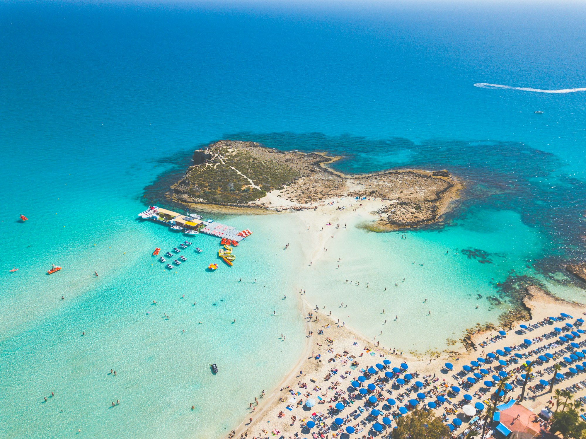 aerial view of packed beach with umbrellas and small island and reef connected by a sandbar