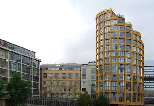 A modern urban scene featuring several buildings with varying architectural styles. On the right, a prominent cylindrical building with yellow framing and large windows stands out. Adjacent to it are rectangular buildings with glass facades, exhibiting a more contemporary design. The sky is overcast, creating a soft, neutral backdrop. Vegetation in the foreground adds a touch of greenery to the urban setting.