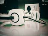 A pair of white headphones with black accents is placed on a reflective surface, with an electrical outlet and other blurred office equipment in the background.