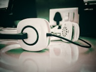 A pair of white headphones with black accents is placed on a reflective surface, with an electrical outlet and other blurred office equipment in the background.