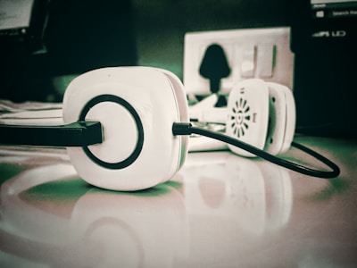 A pair of white headphones with black accents is placed on a reflective surface, with an electrical outlet and other blurred office equipment in the background.