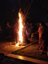 A large bonfire burns brightly, surrounded by people who appear to be participating in a ritual or celebration. The flames illuminate the scene with warm light, contrasting against the dark night. A woman in traditional attire is pouring liquid, possibly part of a ceremonial act.
