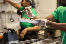 A person wearing a green shirt and gloves is serving food into a tray, using a ladle to pour sauce or gravy. The background includes another person in a green shirt, kitchen equipment, and a tiled wall.