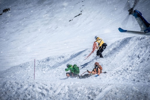 Three individuals in winter gear are situated on a snow-covered slope, with one person kneeling and another holding a coil of rope. Above them, a helicopter is partially visible, indicating a scene of rescue or search operation amidst falling snow.