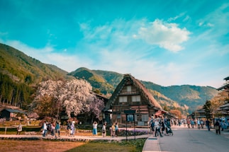 A scenic view of a traditional village nestled in the mountains. There are houses with steep thatched roofs surrounded by greenery and blooming trees. A group of people is gathered on the path in front of the houses, some walking and others standing, suggesting a community or tourist setting. The sky is clear and blue with a few clouds, providing a bright and peaceful atmosphere.