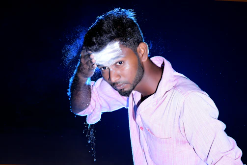 A man in a light pink shirt is shown applying white powder to his forehead. The background is dark, enhancing the contrast with the brightly lit subject. The powder creates a misty effect around his hand.
