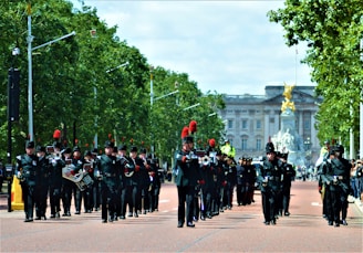 A marching band parades along a wide street lined with trees, with a large, ornate building visible in the background. The musicians wear dark uniforms with red accents and play various instruments.