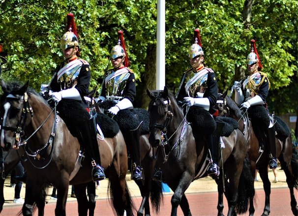 Four equestrians dressed in ornate uniforms ride black horses along a tree-lined avenue. The riders wear reflective helmets with red plumes and carry swords, and their attire is adorned with gold and white details. The scene suggests a formal or ceremonial event, and the trees in the background provide a natural backdrop.