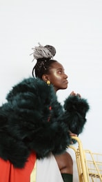 A person with braided hair and metallic accents in their makeup is wearing a luxurious dark fur coat. They are holding onto a yellow metallic frame, possibly part of a cart or chair, and the background is minimalistic and bright, enhancing the contrast with their outfit.