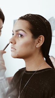 A person is having makeup applied to their face, focusing on the eye area. They have pink and gold tones on the eyelids, neatly tied back hair with decorative elements, and wear a simple earring and necklace.