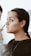 A person is having makeup applied to their face, focusing on the eye area. They have pink and gold tones on the eyelids, neatly tied back hair with decorative elements, and wear a simple earring and necklace.