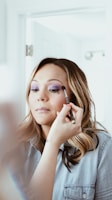 A woman with long, wavy hair is having makeup applied to her face. She is wearing a denim shirt and appears to be in a well-lit room. A hand holding a makeup brush is applying eyeshadow, and her eyes are closed.