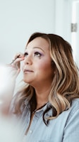 A person with long, wavy hair is receiving makeup, being applied with a brush around the eye area. The setting is bright with natural lighting, and a slight smile is present, suggesting a calm and pleasant atmosphere.
