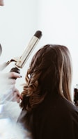 A person is having their hair styled with a curling iron, creating loose curls. The hair is medium length and a light brown shade. The surrounding environment is bright, and there's a soft focus effect on the tools and stylist's hands.