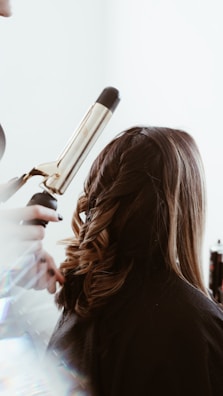 A person is having their hair styled with a curling iron, creating loose curls. The hair is medium length and a light brown shade. The surrounding environment is bright, and there's a soft focus effect on the tools and stylist's hands.