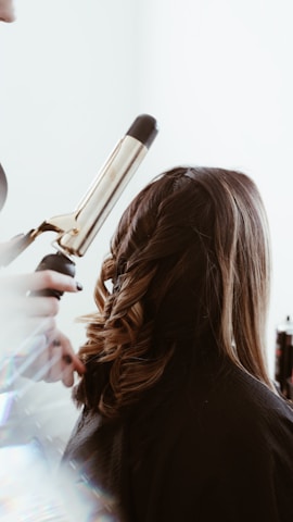 A person is having their hair styled with a curling iron, creating loose curls. The hair is medium length and a light brown shade. The surrounding environment is bright, and there's a soft focus effect on the tools and stylist's hands.