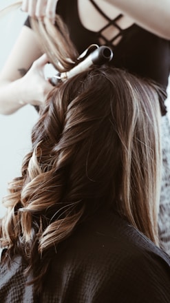 A person is having their hair styled with a curling iron. The image focuses on long, wavy hair with rich brown tones and highlights, with the stylist's hands working on creating curls. The individual is wearing a black cape, suggesting a professional hairdressing setting.